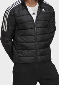 Personne portant une veste puffer noire Adidas avec des rayures blanches sur les manches et le logo Adidas, associée à un pantalon noir et un t-shirt blanc Adidas.