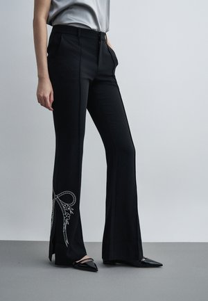 Žena nosiace čierne rozšírené nohavice s bielym vyšitým detailom mašle pri členku a čierne špicaté topánky, stojaca pred jednoduchým pozadím.