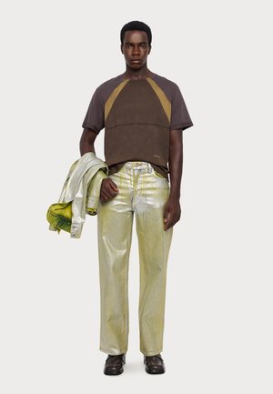 Brun og olivenfarvet farveblokeret t-shirt parret med metallisk gule jeans. Modellen holder en sølvjakke med grøn foring. Sorte sko fuldender looket.