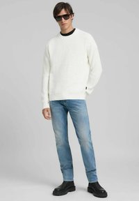 Maglione bianco a coste, colletto interno nero, jeans azzurri, stivali neri robusti e occhiali da sole scuri. Design e vestibilità semplici e moderni.