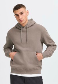 A young man stands against a light background, wearing a beige hoodie with a kangaroo pocket and holding his hands inside the pockets.