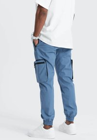 Jogging cargo bleu avec des poignets élastiques, poches latérales et accents de zip noir. Fabriqué en tissu léger, avec une coupe décontractée.