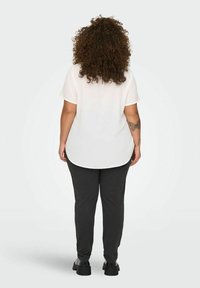 White short-sleeve blouse with a rounded hem, paired with dark gray pants and black footwear. Curly hair and a visible tattoo on the arm.
