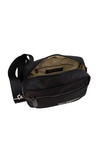 Bolso cruzado de nylon negro con cierre con cremallera, correa ajustable y forro interior beige. Presenta un bolsillo frontal y un detalle de marca.