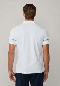 Maglietta polo bianca con colletto, maniche corte e strisce navy ai polsini, realizzata in tessuto morbido. Indossata da una persona vista di spalle.