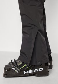 Melni slēpošanas zābaki ar glancētu apdari, ar zaļu akcentu, regulējamiem klikšķiem un plašu pamatni, apvienoti ar melniem slēpošanas biksēm ar rāvējslēdzēju.