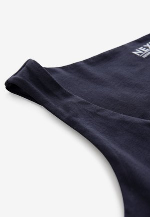 Détail du tissu d’une chemise sans manches bleu marine montrant l’emmanchure et le logo partiel de la marque « NEXT » sur fond blanc.