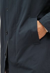 Marineblå jakke lavet af glat stof, med en frontlynlås, store sorte knapper og en sidelomme integreret i designet.
