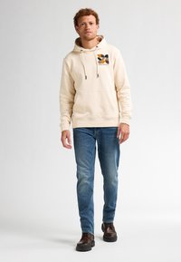 Sudadera con capucha beige y bolsillo frontal, que presenta un diseño gráfico multicolor en el pecho. Combinada con unos vaqueros azules y botas marrones.