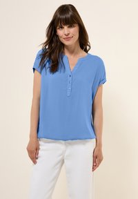 Blusa blu a maniche corte con scollo a V e tre bottoni sul davanti, realizzata in tessuto liscio. Indossata con pantaloni bianchi.