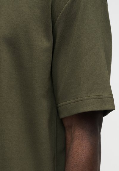 Κοντινό πλάνο του χεριού ενός ατόμου που φοράει πουκάμισο με κοντά μανίκια σε ελιά πράσινο χρώμα, με ορατή υφή υφάσματος και ραμμένο τελείωμα στα μανίκια.