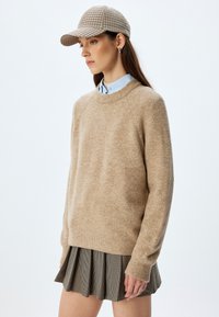 Beige Strickpullover mit Rundhalsausschnitt, kombiniert mit einer karierten Mütze und einem plissierten Schwarz-Weiß-Rock. Glatte Textur, lockere Passform.