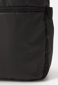 Sac à dos noir en tissu lisse et texturé, avec un logo imprimé sur le devant. Comprend des bretelles en toile et un compartiment zippé.