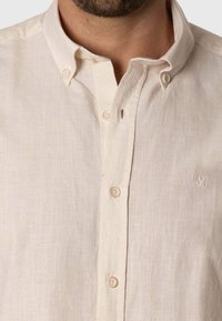 Camisa de lino color crema con botones, que presenta un cuello clásico, una textura sutil y un logo bordado en el lado izquierdo del pecho.