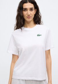 Camiseta blanca de algodón con mangas cortas, que presenta un logo verde bordado en el pecho. Corte recto y cuello redondo.
