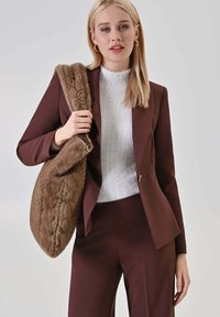 Blazer marrone sartoriale indossato sopra un maglione grigio chiaro con trama, abbinato a pantaloni marroni, con una grande borsa marrone pelosa a tracolla.