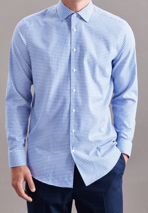 Homme portant une chemise à manches longues à petits carreaux bleus clairs et blancs avec un pantalon foncé, main dans la poche sur fond gris.