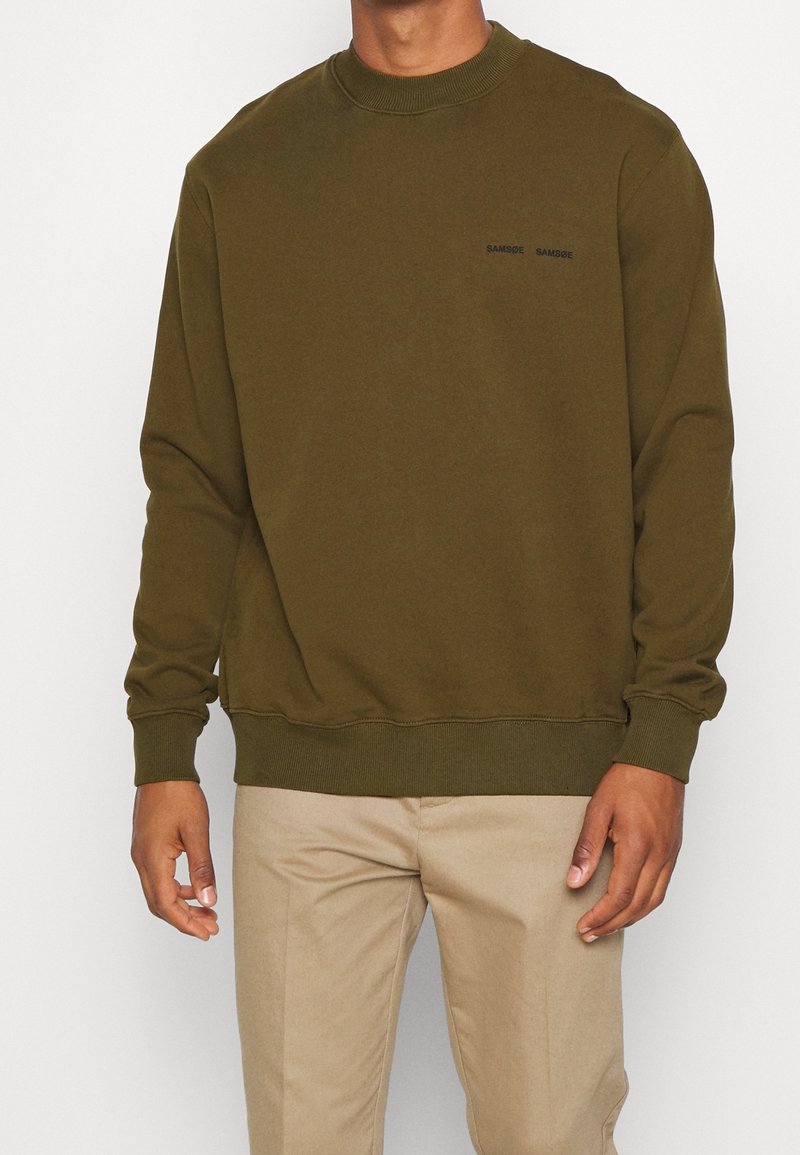 Sweatshirt vert olive à col rond, avec des poignets et un bas côtelés. Présente un petit logo noir sur la poitrine. Texture de tissu douce.