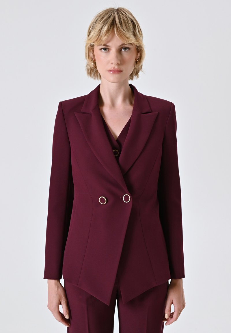 Femme blonde portant un blazer bourgogne ajusté à double boutonnage avec des boutons circulaires dorés, se tenant devant un fond clair uni.