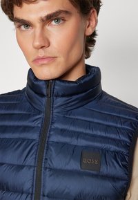 Gilet imbottito blu navy con colletto alto, caratterizzato da cuciture orizzontali e una patch nera con logo. Chiusura con zip e texture elegante.