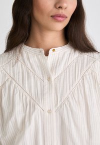 Light-colored, striped blouse with a round neckline, button closure, and pleated details. Features a golden button accent and delicate stitching.