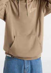 Sweat à capuche beige avec une poche avant, arborant un logo "Vans" clair sur la poitrine. Tissu doux avec une coupe décontractée.