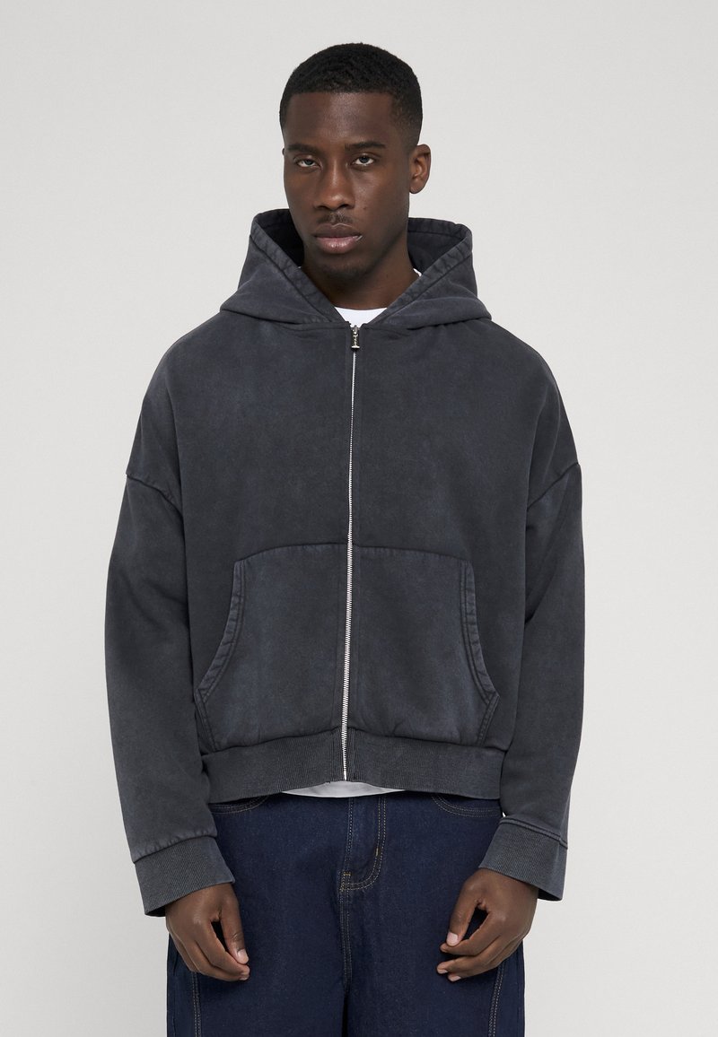 Jeune homme portant un sweat à capuche gris foncé zippé avec des poches avant et un jean bleu, debout devant un fond clair uni.