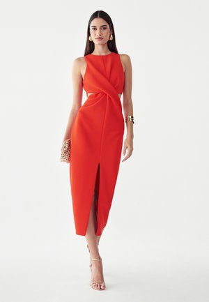 Vrouw draagt een rode mouwloze midijurk met een voorsplit, houdt een kleine gestructureerde clutch vast en draagt gouden oorringen en een bangle armband.