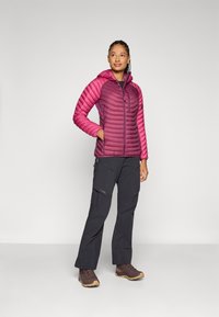 Chaqueta aislante ligera, con capucha, de color rosa y diseño acolchado; combinada con pantalones negros y calzado marrón, en un fondo neutro.