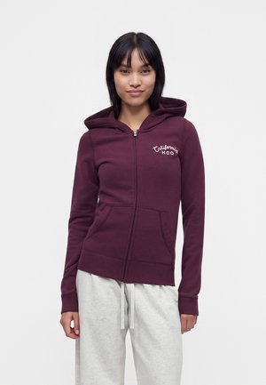 Frau trägt einen bordeauxroten Hoodie mit Reißverschluss und dem Text "California HCO" auf der Brust und hellgraue Jogginghosen, sie steht vor einem einfarbigen Hintergrund.