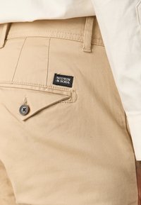 Beige broek met een achterzak met een knoopsluiting en een zwart "Scotch & Soda" label, gecombineerd met een zichtbaar wit manchetje van een shirt.