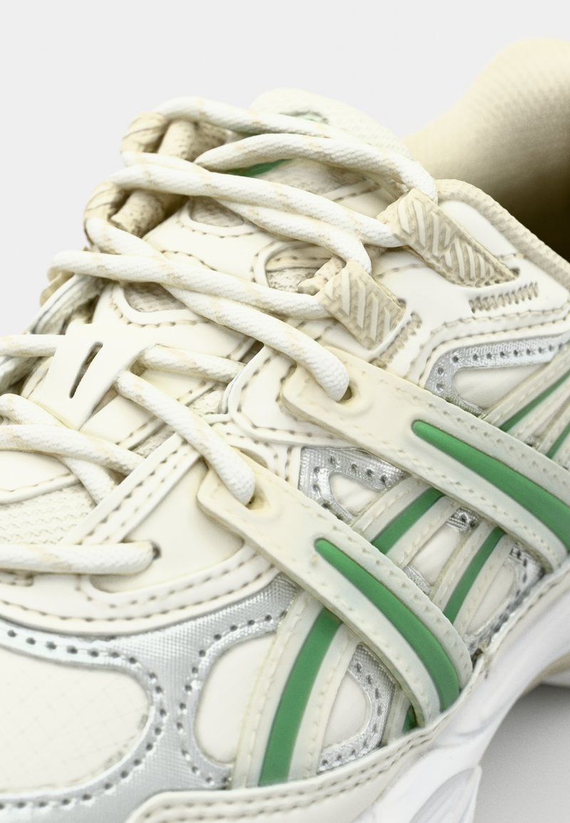 Chaussure de sport blanche en matériau synthétique et en maille, avec des accents verts et des lacets texturés. La vue en gros plan met en évidence les détails.