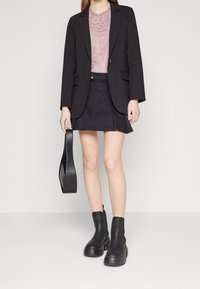 Femme portant un blazer noir, une mini-jupe noire, un top en dentelle rose, des bottines noires, tenant un sac à main noir sur fond uni.