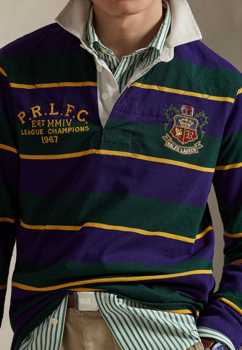 Maillot de rugby rayé violet et vert avec des rayures jaunes, un blason brodé et un col blanc, superposé à une chemise rayée verte et blanche.