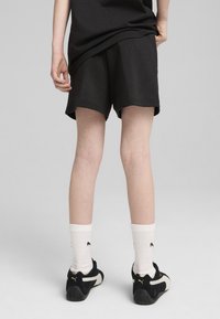 Pantalones cortos deportivos negros con una textura suave, combinados con calcetines blancos acanalados que tienen un logo negro, y zapatillas negras con detalles dorados.
