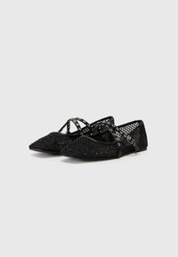 Ballerines noires avec un bout pointu, un tissu texturé avec un motif perlé, et des lanières entrecroisées ornées d'accents en matériel décoratif.