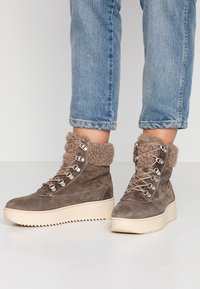 Bottines en suédine marron avec des revers en taupe duveteux, dotées d'œillets en métal et de lacets, sur une semelle en plateforme beige avec un motif en zigzag.