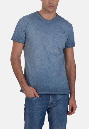 Uomo che indossa una T-shirt a maniche corte blu sbiadito con una piccola tasca sul petto e jeans blu, in piedi con una mano nella tasca.