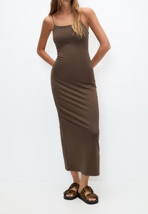 Vrouw in een aansluitende bruine mouwloze midi-jurk met spaghetti bandjes en sandalen met luipaardprint, staand tegen een witte achtergrond.