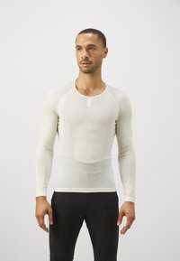 Leichtes, langärmliges Oberteil in Beige mit figurbetontem Schnitt, Rundhalsausschnitt und minimaler Nahtführung. Kombiniert mit schwarzen Sportleggings.