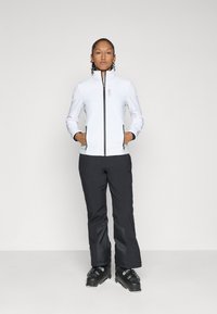 Weiße Softshelljacke mit hohem Kragen, Reißverschluss und zwei vorderen Taschen, kombiniert mit schwarzen Skihosen und schwarzen Skistiefeln.