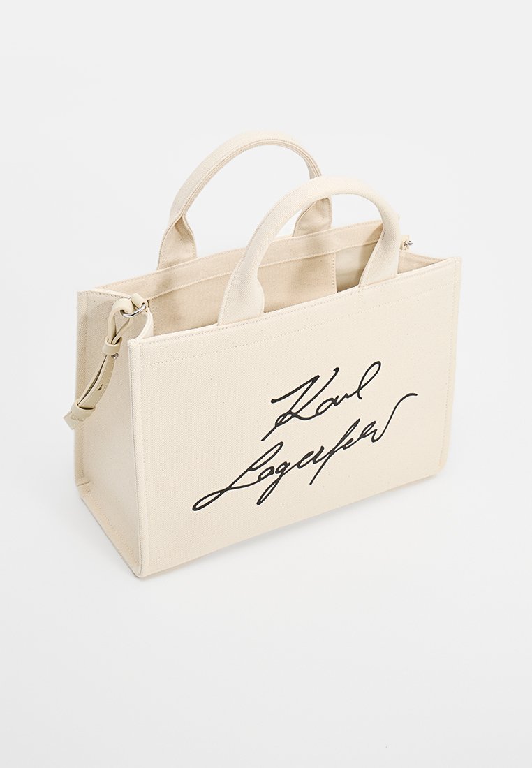 Sac fourre-tout beige avec double poignées, forme rectangulaire, et logo noir signature à l'avant. Fabriqué en toile robuste.