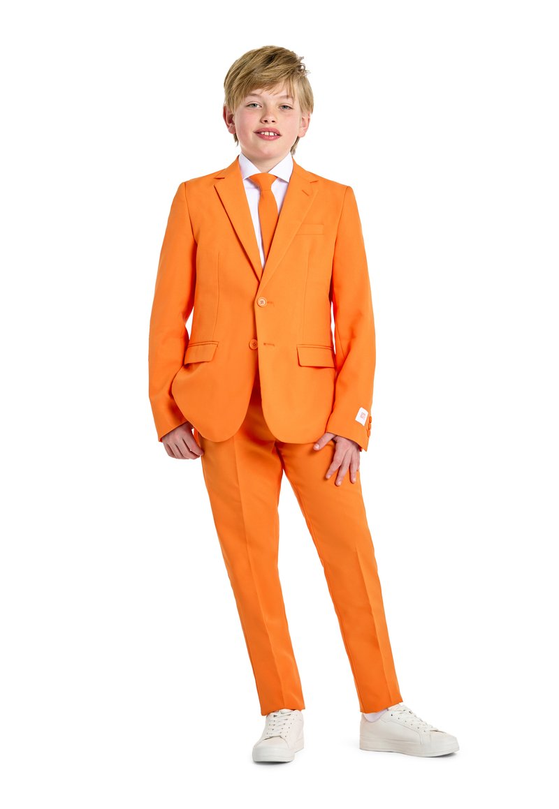 Completo arancione con giacca a un bottone e cravatta abbinata. I pantaloni hanno un taglio dritto. Indossato con sneakers bianche. Tessuto liscio.