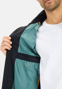 Schwarze Jacke mit einem türkisfarbenen Innenfutter. Mit einem orangefarbenen Akzent am Kragen und einem runden Logo mit der Aufschrift "ULTRA LIGHT." Glatte Textur.