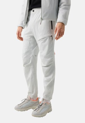 Man die lichtgrijze taps toelopende broek met ritszakken en lichtgrijze sportschoenen met oranje accenten draagt, staand tegen een effen achtergrond.