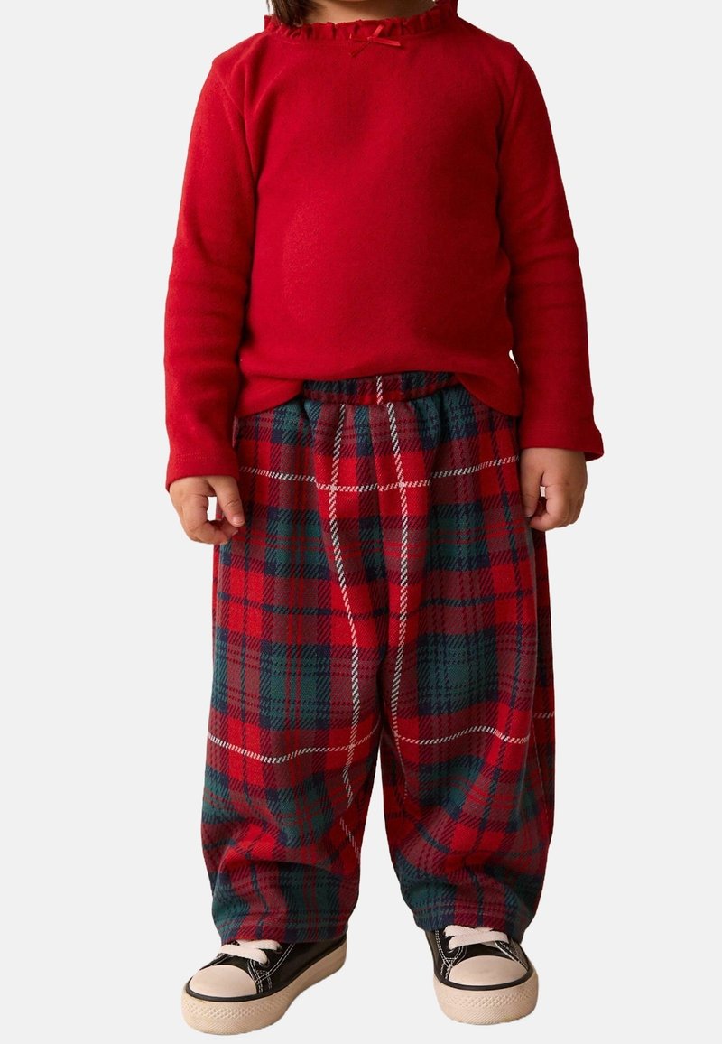 Enfant portant un haut rouge à manches longues, un pantalon ample à carreaux rouge et vert, et des baskets noires avec des semelles et des lacets blancs, debout sur un fond blanc.