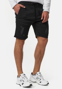 Mustat farkkushortsit kuluneilla yksityiskohdilla ja ribbineuloksilla, joissa on joustava vyötärönauha ja kiristysnauha. Yhdistettynä valkoisiin urheilukenkiin.