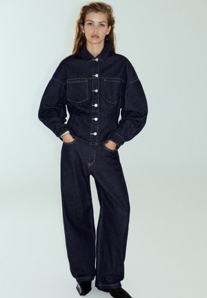 Jeune femme debout, les mains dans les poches, portant une veste en jean foncée et un jean à jambes larges avec des chaussures noires pointues sur fond uni.