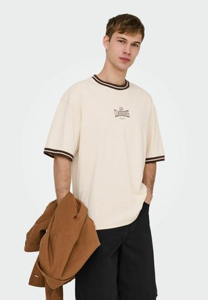 Młody mężczyzna w beżowej koszulce z brązowym obszyciem i napisem, trzymający brązową kurtkę w jednej ręce, w czarnych szortach, z ręką w kieszeni.