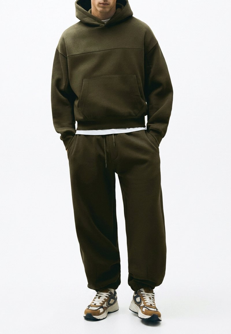 Homme debout les mains dans les poches, portant un sweat à capuche vert foncé, un pantalon de survêtement assorti, un t-shirt blanc et des baskets multicolores.